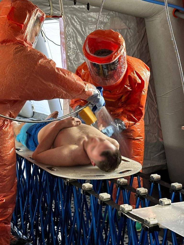 Eine liegende Person wird durch Einsatzkräfte in Schutzkleidung abgeduscht.