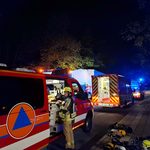 Nächtliche Einsatzstelle: Eine Einsatzkraft mit Helm steht am geöffneten Geräteraum eines Fahrzeugs mit Katastrophenschutz-Zeichen. Dahinter weitere Einsatzfahrzeuge mit Blaulicht, auf dem Gehweg liegen Atemschutzgeräte.